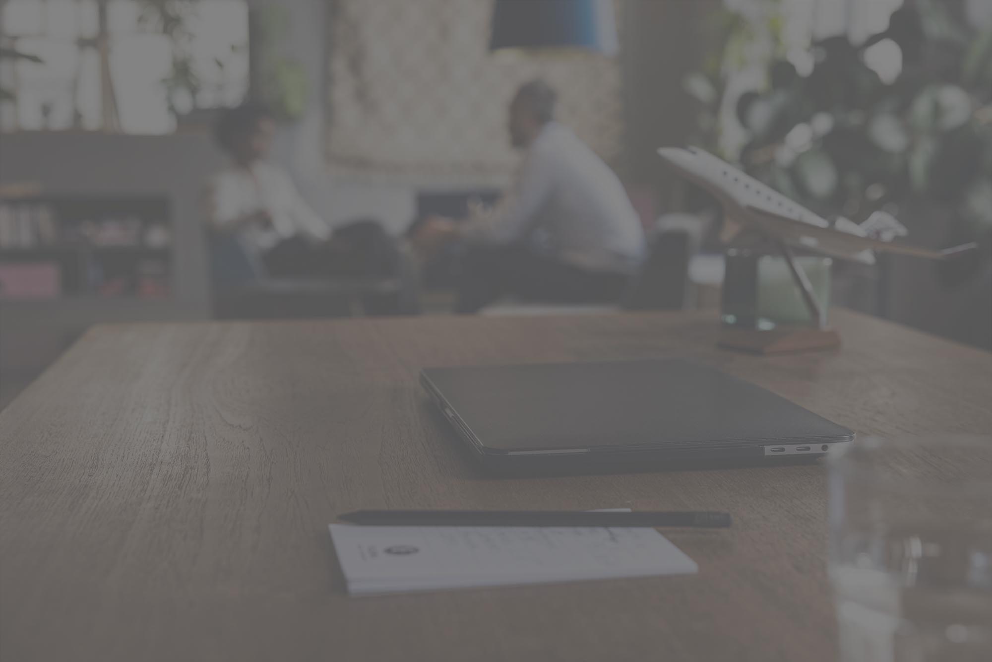 Image with grey overlay of wooden desk, a black laptop and model airplane in focus. A meeting is in the blurred background